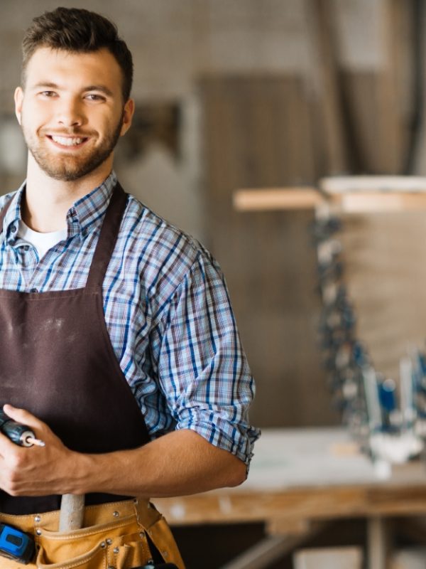 Waist-up portrait of smiling bearded craftsman with electric drill in hands standing in spacious workshop and looking at camera