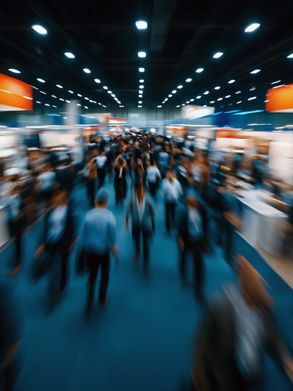 A blurred image of a convention hall filled with people. The scene features a blue floor, orange signs, and a dark ceiling with lights. People in suits and shirts are walking.