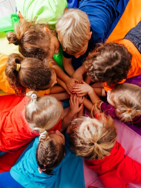 Lovely children laying on multicolor canopy in circle. Team building game for daycare children in colorful t-shirts. Top view kids on wooden floor.